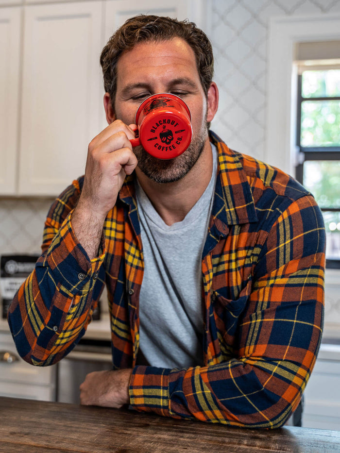 Man in a plaid shirt drinking from a red mug in a kitchen.